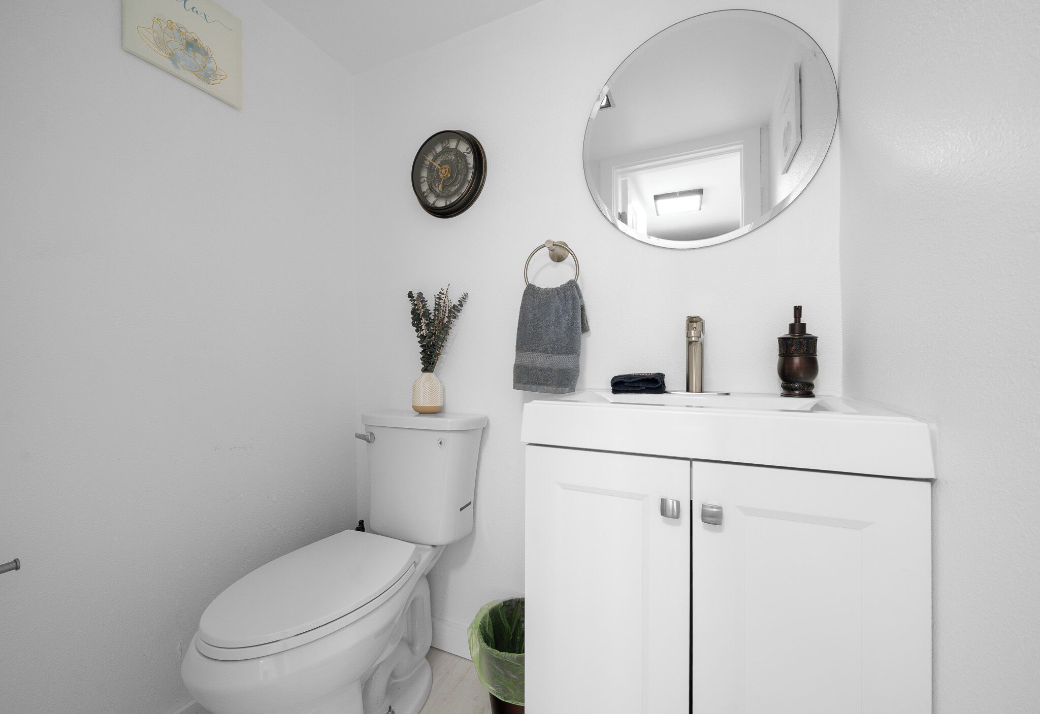 A white bathroom with a toilet, a sink with a mirror above it, a clock, and a trash bin.