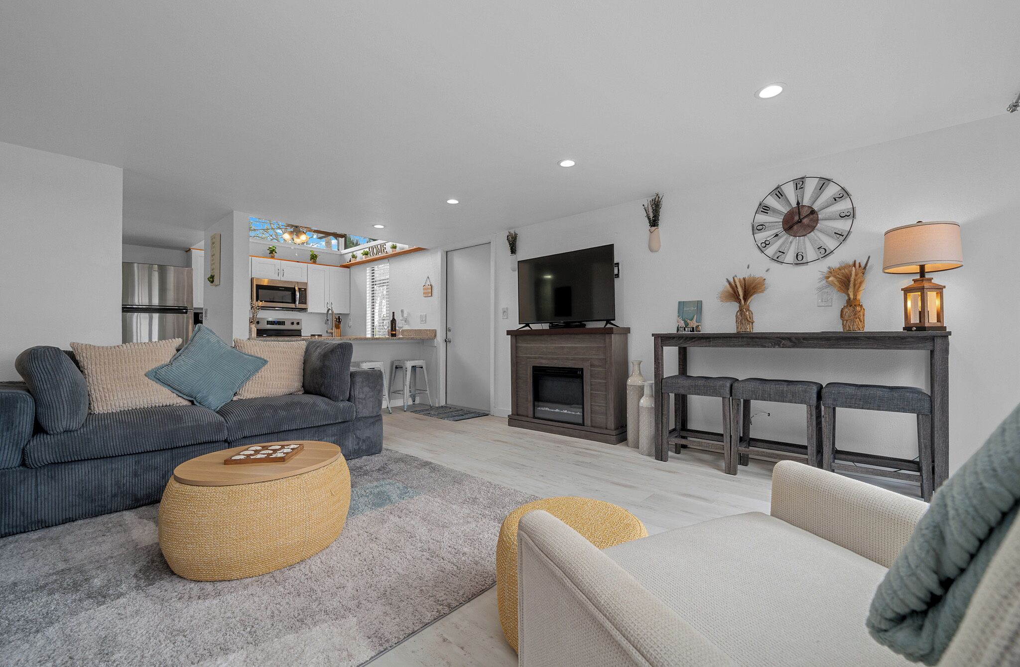 Una sala moderna con un sofá gris, una mesa de centro redonda y una chimenea. Se ve una cocina al fondo, con armarios blancos y un refrigerador de acero inoxidable.