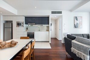 A modern kitchen with white cabinets, a microwave, and an oven. There is a dining table with chairs and a sofa in the living area. - Deluxe Two Bedroom Hilton Apartment (Surfers Paradise)