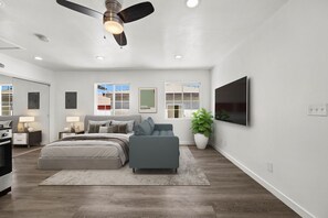 A modern bedroom with a bed, a sofa, a ceiling fan, a TV, and a large window. - Bright & Modern Bixby Knolls Studio + In-Unit W/D (Long Beach)