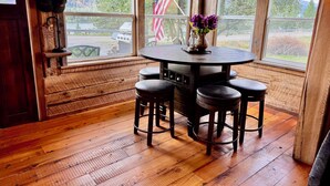 Een houten interieur met een ronde tafel en vier krukken, grote ramen met een Amerikaanse vlag, en een uitzicht op een meer en bergen.