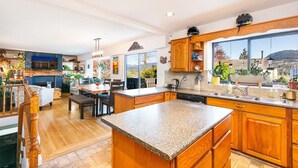 Una cocina amplia con armarios de madera, encimera de granito y una isla grande. Hay un comedor con mesa y sillas, y vistas al exterior a través de ventanas grandes.