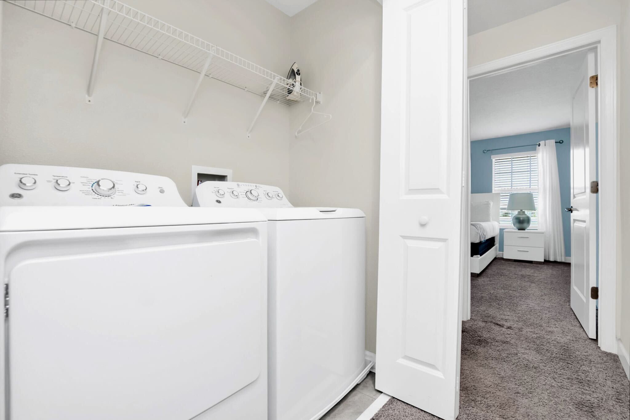 A laundry area with two white washing machines, a drying rack, and a door leading to a bedroom with a bed, nightstand, and a window with curtains.