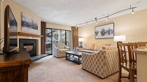 A spacious living room with a fireplace, comfortable seating, and large windows offering a view of trees. - Walk to Vail Village 1BR Condo w/ Free Shuttle to the Slopes (Vail)