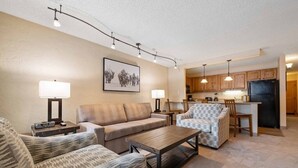 A living room with a sofa, armchairs, a coffee table, and a dining area with a chair and table. There is a kitchen in the background with wooden cabinets and a refrigerator. - Walk to Vail Village 1BR Condo w/ Free Shuttle to the Slopes (Vail)