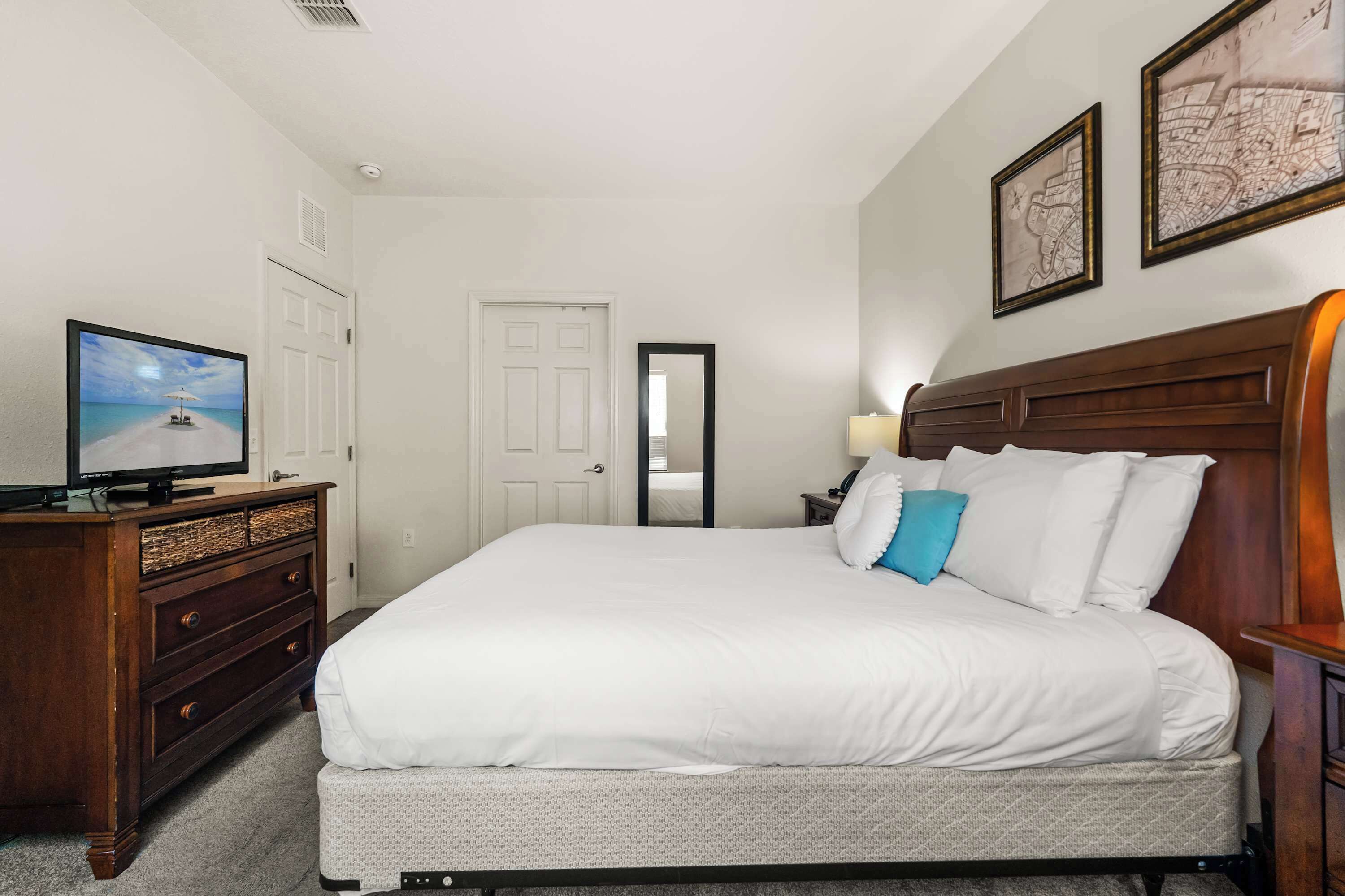 A hotel room with a bed, a television on a dresser, a mirror, and two framed maps on the wall.