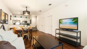 A modern living room with a flat-screen TV, a dining area with chairs, a kitchen with white cabinets, and a staircase leading to another floor. - Upstay Modern 3BR Townhome With Pool Near Disney (Kissimmee)
