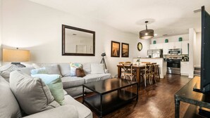 A modern living room with a sofa, coffee table, dining table, and kitchen area with appliances. - Upstay Modern 3BR Townhome With Pool Near Disney (Kissimmee)