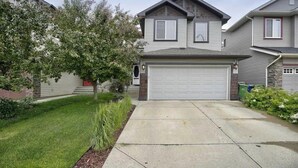 A two-story residential house with a front garage, a well-maintained lawn, and a large tree in the front yard. - Charming Basement Suite With Parking & Laundry (Calgary)