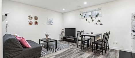 A modern living room with a grey sofa, a coffee table, a TV, and a dining area with chairs and a table.
