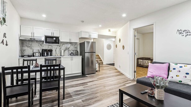 A modern kitchen with white cabinets, a stainless steel refrigerator, and a microwave. There is a dining area with a table and chairs, and a living room with a sofa and a coffee table. - Charming Basement Suite With Parking & Laundry (Calgary)
