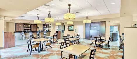 A spacious dining area with multiple tables and chairs, a kitchen in the background, and pendant lighting.