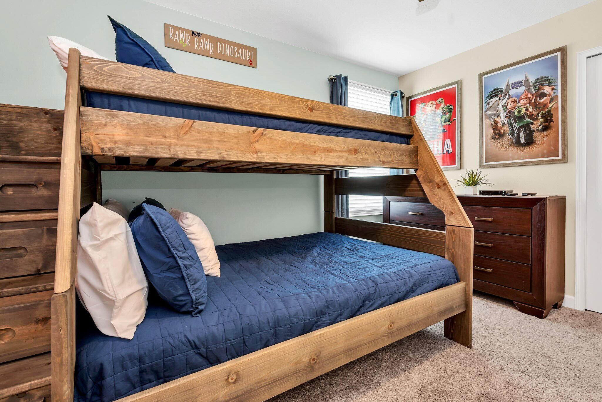 Un lit superposé avec une structure en bois, un matelas bleu et des oreillers. Il y a une commode en bois sur la droite et une affiche accrochée au mur.