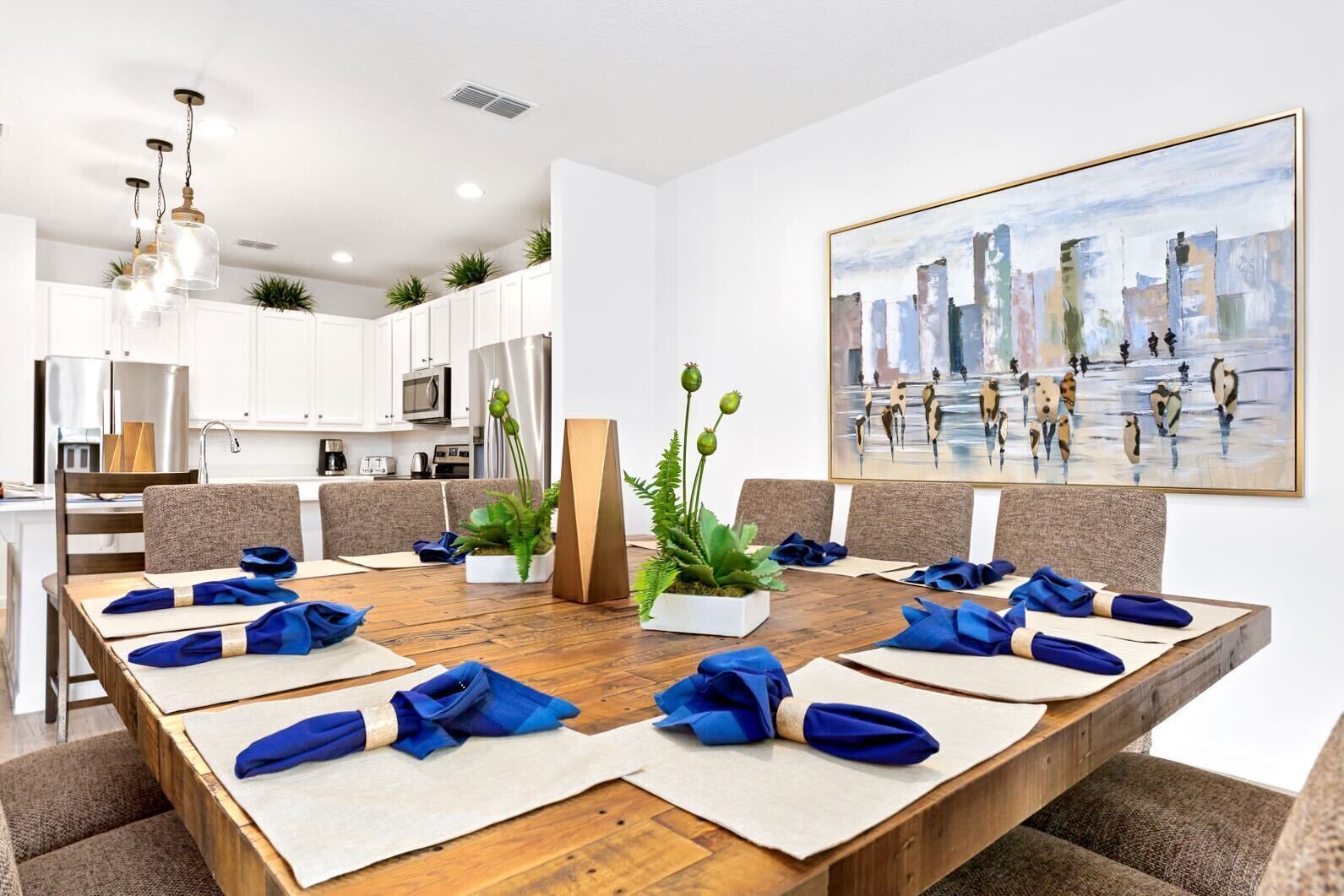 A modern dining area with a long wooden table set for a meeting, surrounded by chairs, with a cityscape painting on the wall and a kitchen in the background.