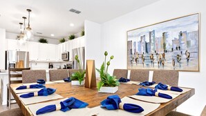 A modern dining area with a long wooden table set for a meeting, surrounded by chairs, with a cityscape painting on the wall and a kitchen in the background. - Upstay: Luxury 10br Mansion w Pool,hot Tub & Games (Kissimmee)