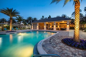 Un espace piscine doté d’un patio couvert, de palmiers et d’un coin salon bien éclairé.