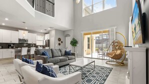 A spacious living area with a gray sofa, white armchairs, and a patterned rug. A dining area with a table and chairs is visible in the background. - Upstay Modern 5BR Resort Home w Pool & Hot Tub (Kissimmee)