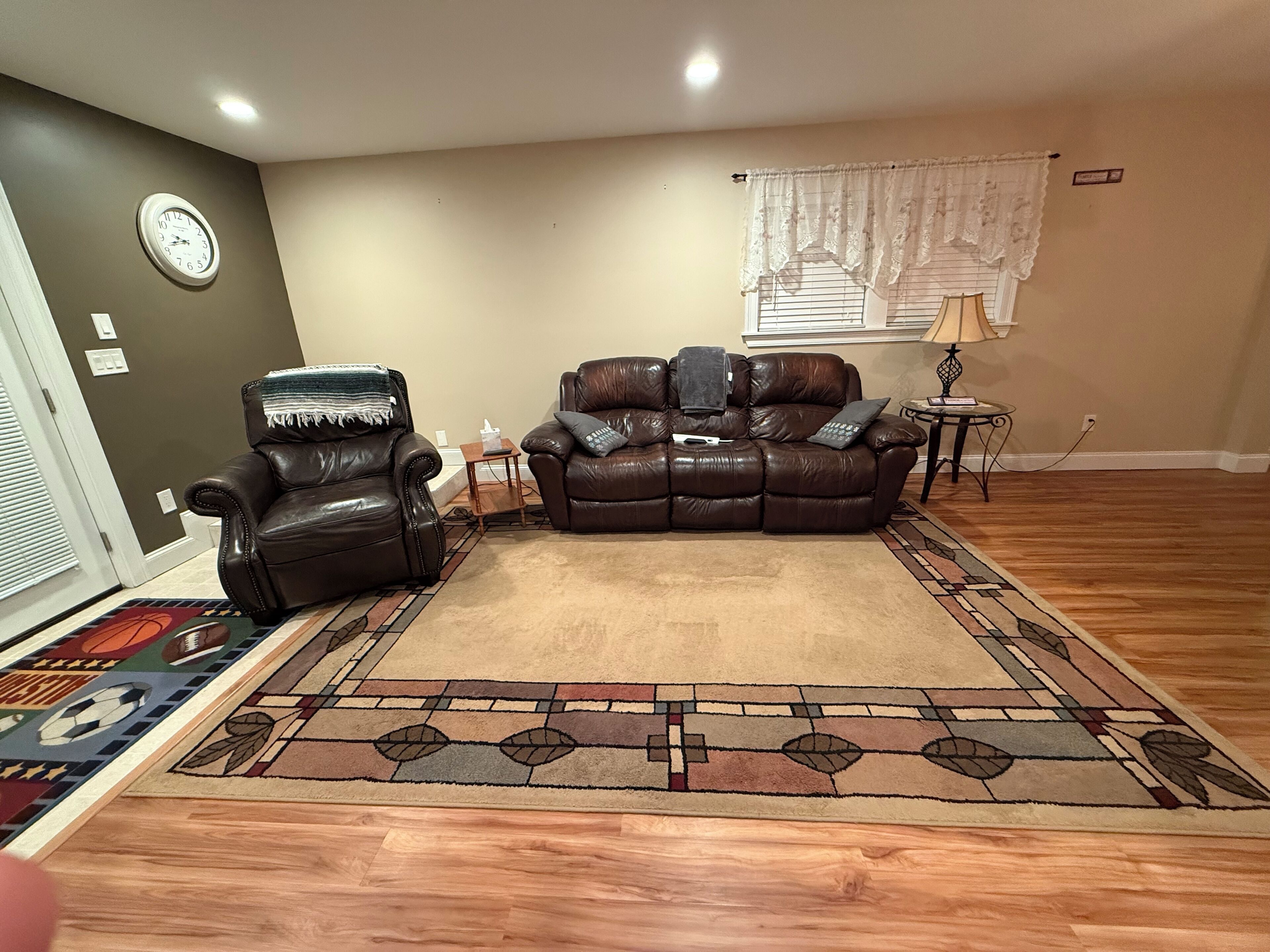 A living room with a brown leather sofa, a leather chair, a patterned rug, a clock on the wall, and a window with a curtain.