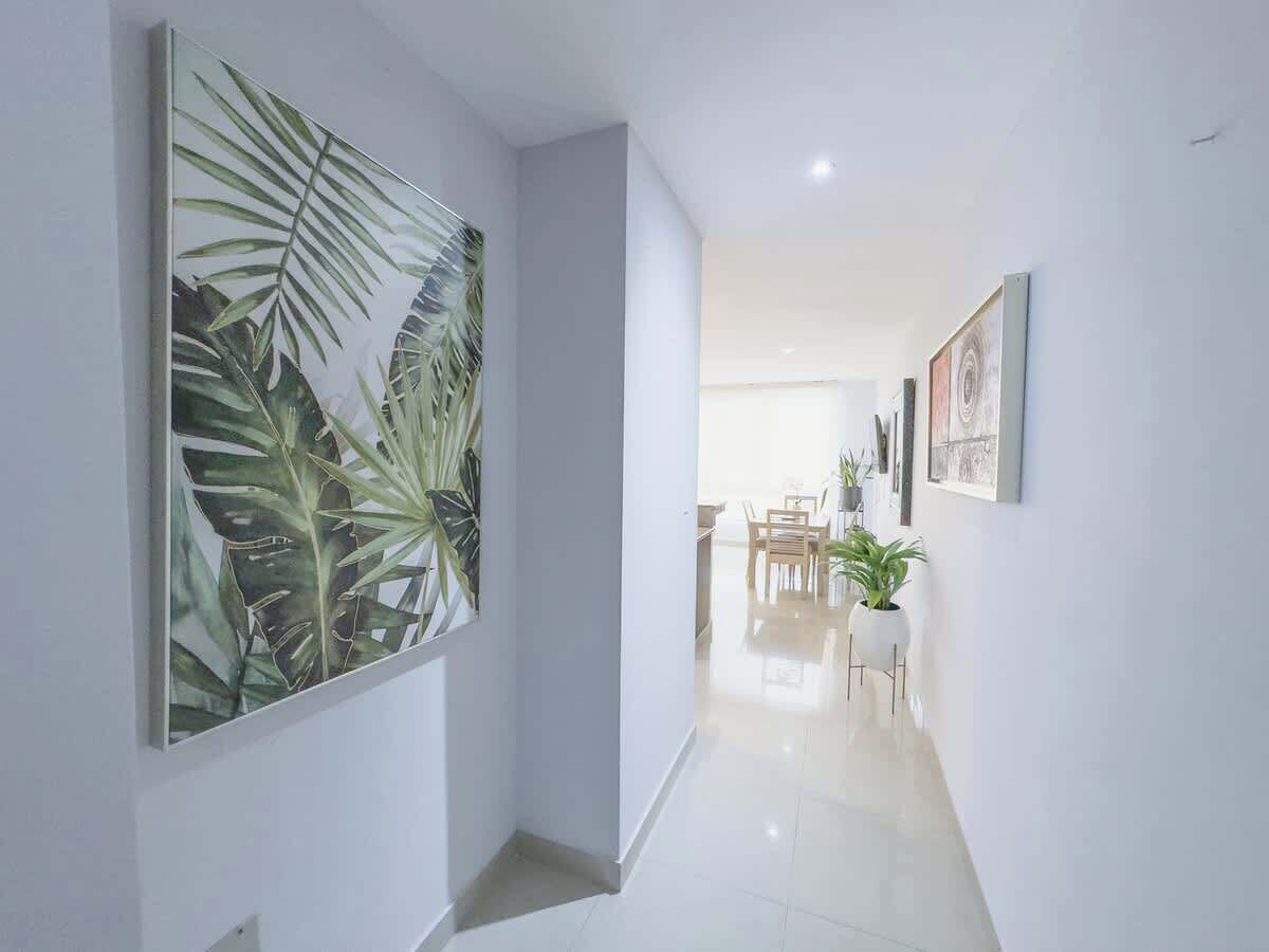 Un pasillo con un cuadro de una hoja tropical grande, una zona de comedor con sillas y una mesa, y una planta en maceta.