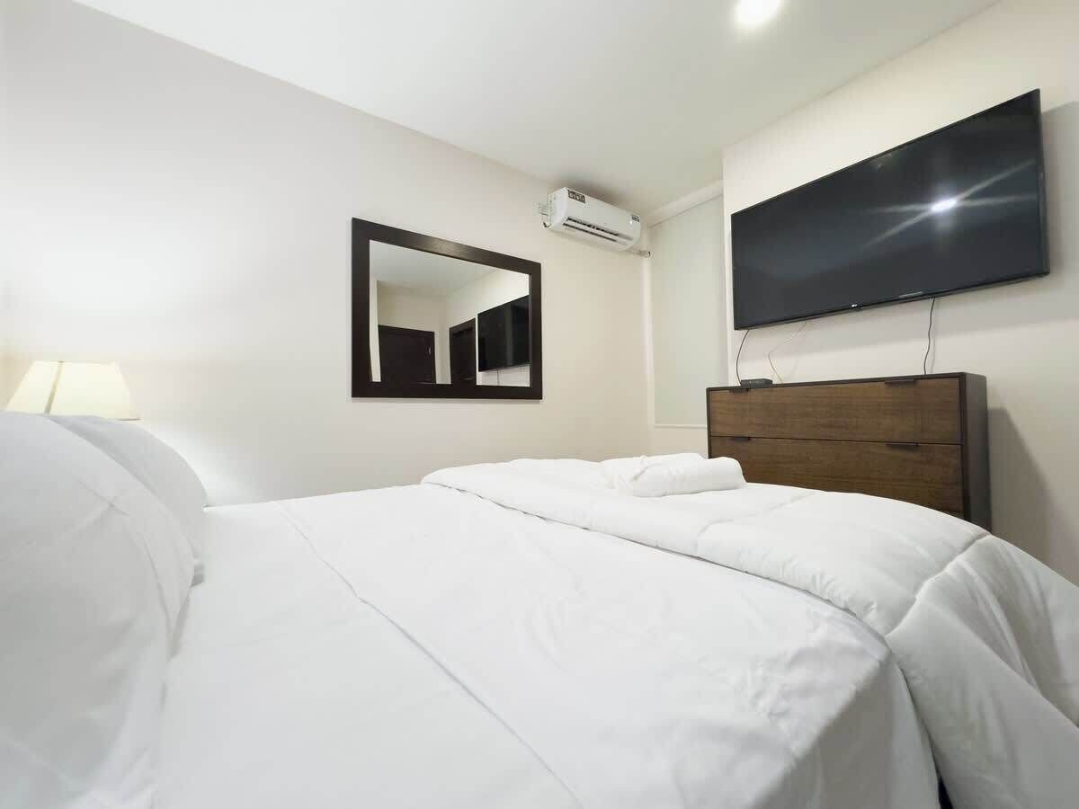 A hotel room with a bed, a flat-screen TV mounted on the wall, and an air conditioning unit.