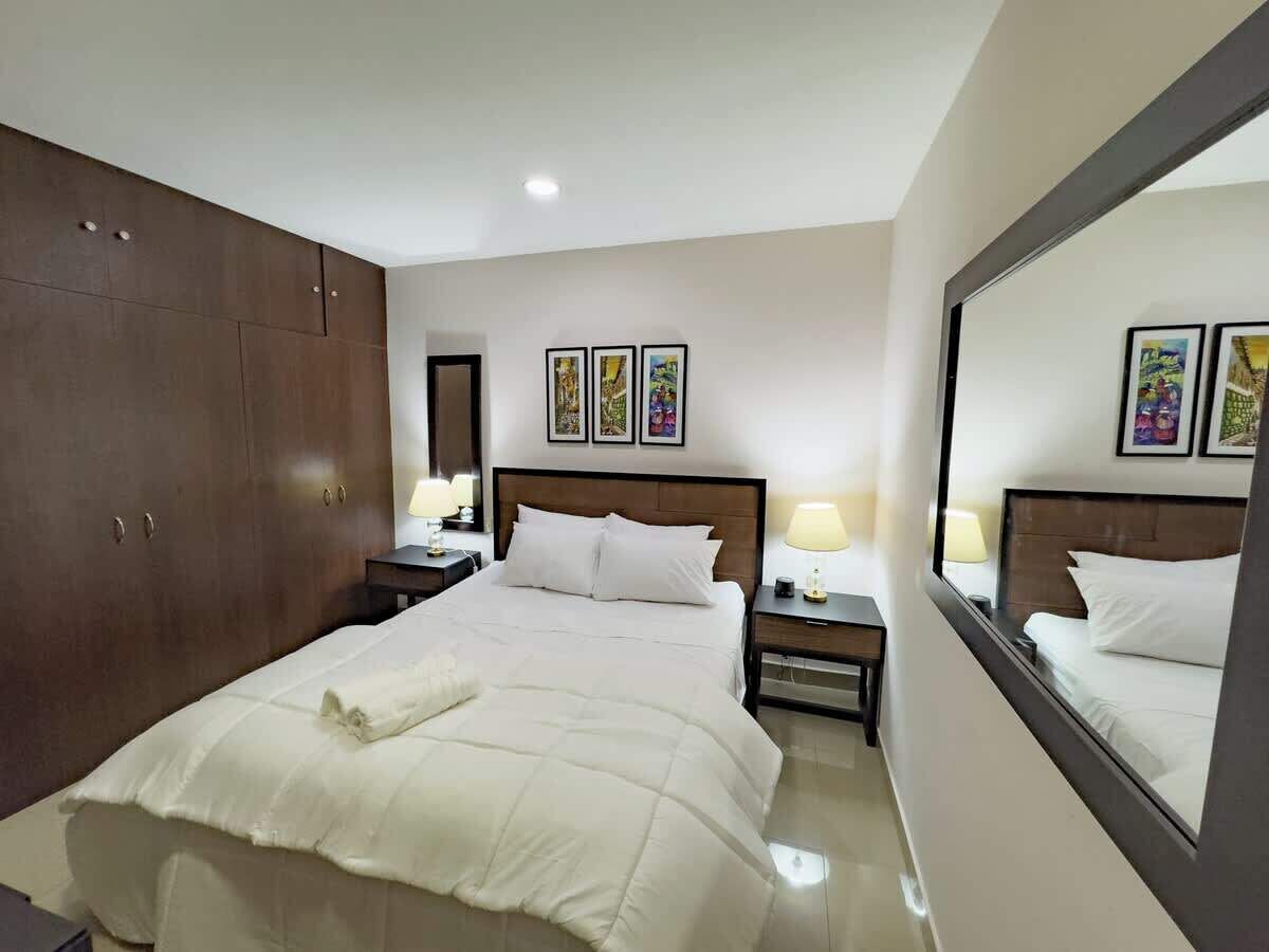 Habitación de hotel con una cama grande, un armario, una mesita de noche con lámpara y un espejo que refleja la cama.
