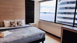 Habitación de hotel moderna con un ventanal grande, cama con almohadones estampados y vista a un cuerpo de agua y edificios.
