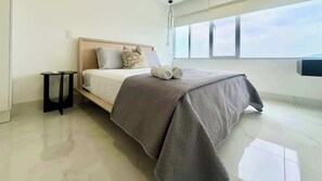 Une chambre d’hôtel moderne, dotée d’un grand lit, d’une petite table de chevet et offrant une vue sur le ciel par une grande fenêtre.