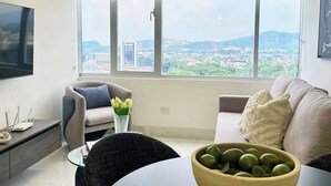 Un salon moderne doté d’une grande fenêtre, d’un téléviseur à écran plat, d’un canapé, d’une table à manger et d’un saladier rempli de fruits.
