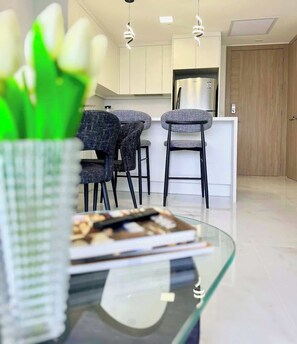 A modern kitchen with white cabinets, a stainless steel refrigerator, and a dining area with a glass table and two chairs.