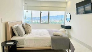 Une chambre moderne dotée d’une grande fenêtre, d’un lit, d’une table de chevet, d’un miroir et d’un téléviseur à écran plat.
