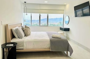 Un dormitorio moderno con un ventanal grande, una cama, una mesita de noche, un espejo y un televisor de pantalla plana.