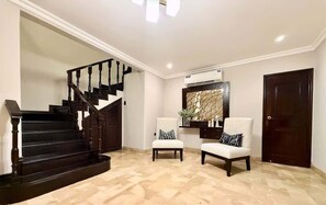 A staircase with dark wooden railings, a wall-mounted air conditioning unit, two upholstered chairs with patterned cushions, and a dark wooden door. - Large Apt in Lomas Urdesa • Sleeps 8 • BBQ (Guayaquil)
