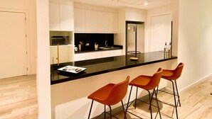 A modern kitchen with a black countertop, white cabinets, and a built-in oven. There are three orange bar stools and a small island with a sink. - Luxurious Escape at Santana lofts includes Parking (Guayaquil)