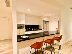 A modern kitchen with a black countertop, white cabinets, and a built-in oven. There are three orange bar stools and a small island with a sink. - Luxurious Escape at Santana lofts includes Parking (Guayaquil)