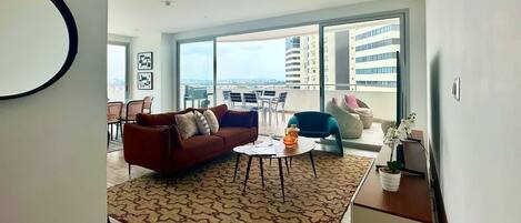 A modern living room with a sofa, coffee table, and chairs, a balcony with a view of buildings, and a large window.