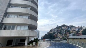 A modern building with a curved facade, a swimming pool with lounge chairs, and a hillside residential area in the background. - Luxurious Escape at Santana lofts includes Parking (Guayaquil)