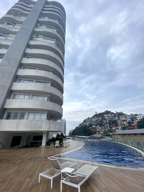 A modern building with a curved facade, a swimming pool with lounge chairs, and a hillside residential area in the background. - Luxurious Escape at Santana lofts includes Parking (Guayaquil)