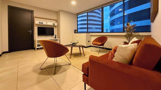 Un soggiorno moderno con un divano, due poltrone, un tavolino, una TV e una grande finestra vista su un edificio.