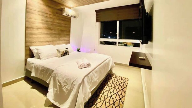 A modern bedroom with a wooden ceiling, a bed with white linens, a nightstand, a TV, and a window with a view of the city at night. - Poolside Condo w/Parking Near US Consulate (Guayaquil)