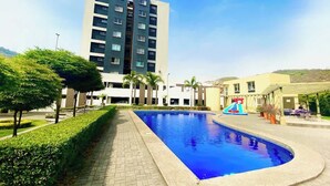 A swimming pool with a slide, surrounded by a landscaped area with hedges and trees, and a multi-story building in the background. - Poolside Condo w/Parking Near US Consulate (Guayaquil)