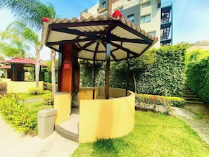 A covered outdoor seating area with a curved bench, a trash bin, and a building in the background. - Amazing 2BD apt near US Consulate includes Parking (Guayaquil)