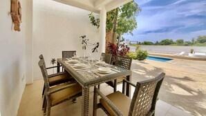 Un comedor con una mesa lista para cuatro personas, rodeado de sillas de mimbre, un ventilador de techo y vistas a una piscina y follaje.