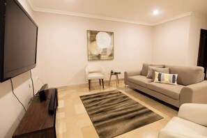 Un salon équipé d’un téléviseur à écran plat, d’un canapé beige, d’un fauteuil blanc, d’une petite table d’appoint et d’un tableau abstrait encadré accroché au mur.