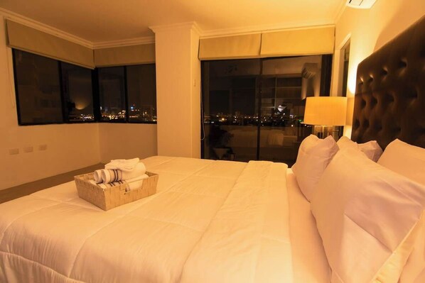 Une chambre d’hôtel avec un grand lit, une table de chevet, une lampe et une vue sur la ville la nuit.