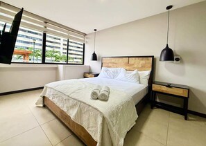 A modern hotel room with a large bed, two bedside tables, a flat-screen TV mounted on the wall, and a window with blinds. - Stylish 3BR Apartment with River View & Nightlife (Guayaquil)