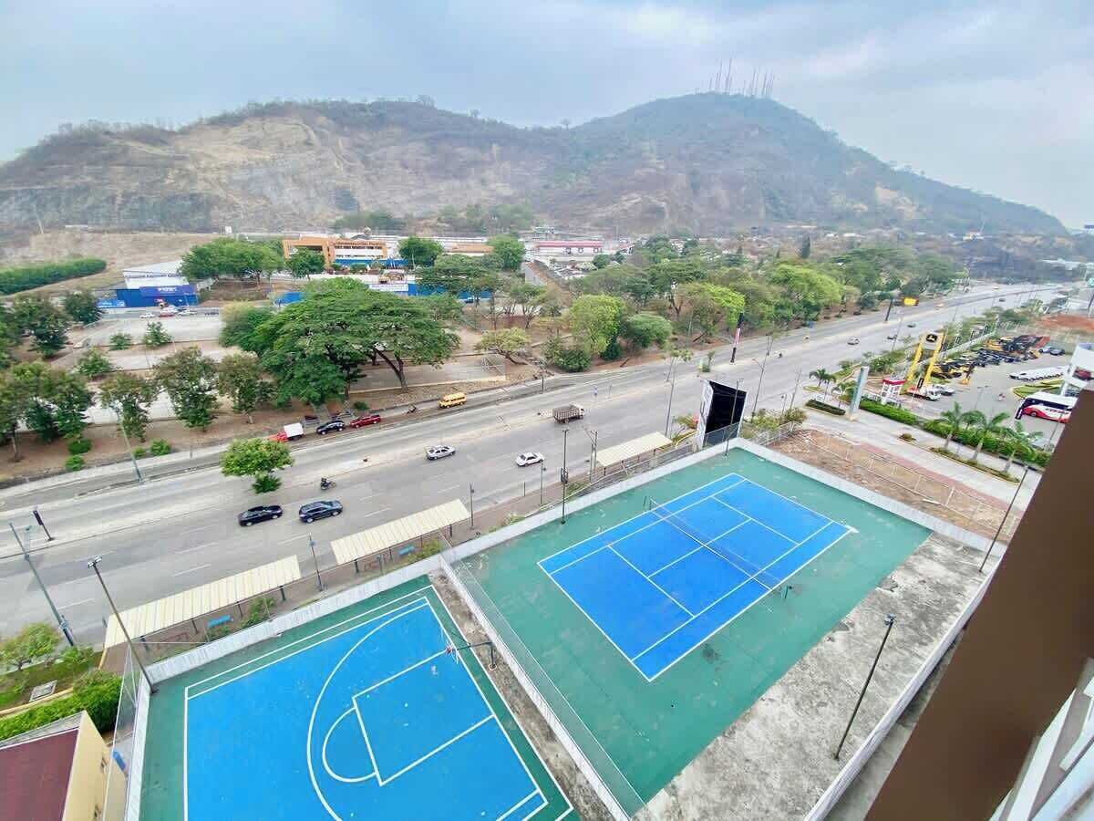 A view of a tennis court complex with multiple courts, a road with vehicles, and a mountainous backdrop.
