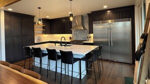 Una cucina moderna con mobili scuri, un'isola centrale con sgabelli da bar, un frigorifero in acciaio inossidabile e illuminazione a sospensione.
