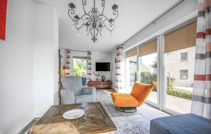 A modern living room with a wooden coffee table, a grey armchair, an orange chair, and a blue sofa. A chandelier hangs from the ceiling, and a flat-screen TV is mounted on the wall. - 3 bedroom beautiful home in Röbel (Röbel)
