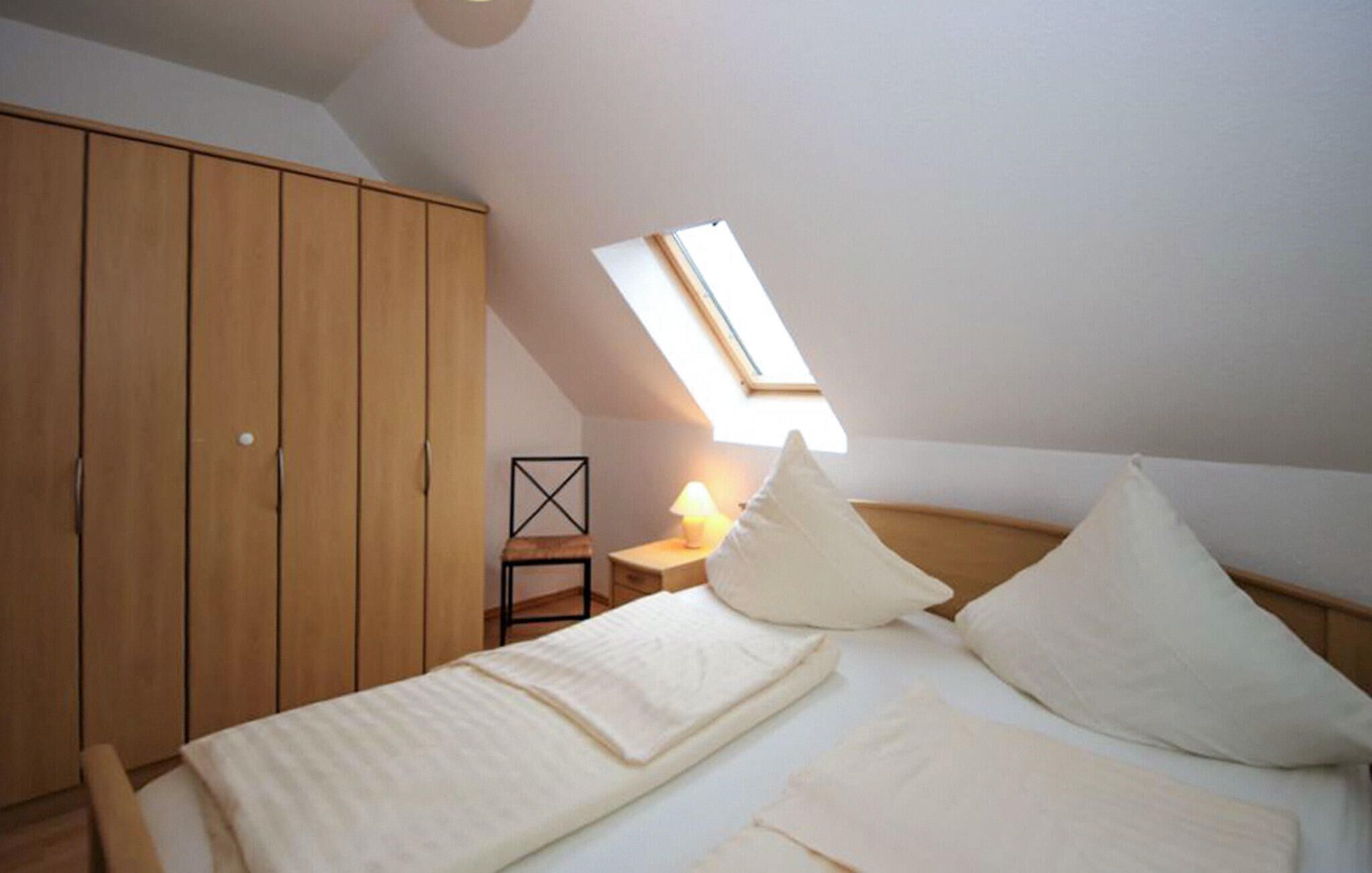 Une chambre avec un plafond en pente, un puits de lumière, un lit avec des oreillers, une table de chevet avec une lampe et une armoire en bois.