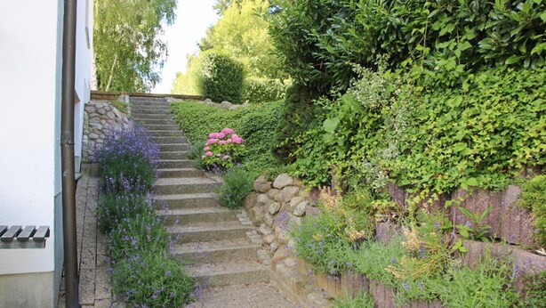 Eine steinerne Treppe führt zu einem Haus, umgeben von üppigem Grün und blühenden Pflanzen.